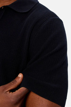 Close-up of a person’s arm and torso wearing the Industrie The Valencia Polo, a short-sleeved black shirt with ribbed texture made from breathable fabric. Their left hand rests on their right arm against a plain light background.