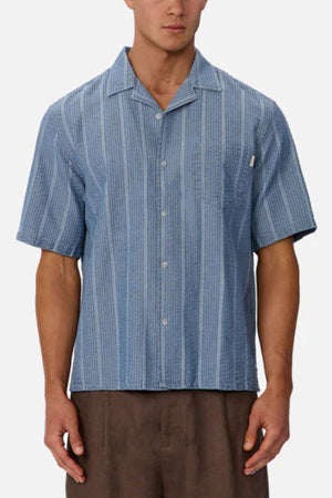 A man wearing the Industrie The Kimridge Short Sleeve Shirt, a blue textured camp collar shirt with vertical stripes, and brown pants stands against a plain light background.