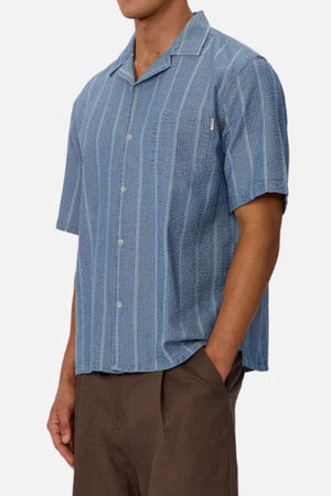 A person wears the Industrie The Kimridge Short Sleeve Shirt, a light blue camp collar shirt with vertical stripes, paired with brown pants. Their hand is in their pocket and their face is not visible.