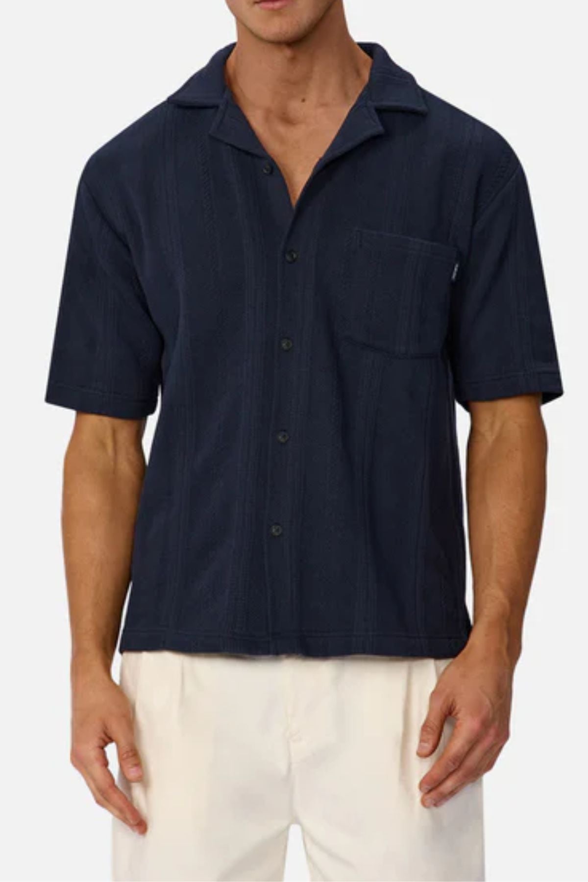 A man wearing an Industrie The Malaga Short Sleeve Shirt in navy blue with a chest pocket and off-white pants, posed against a plain light background.