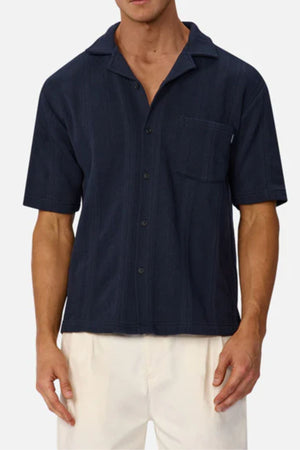 A man wearing an Industrie The Malaga Short Sleeve Shirt in navy blue with a chest pocket and off-white pants, posed against a plain light background.