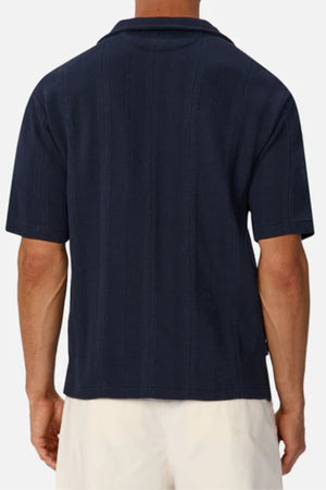 A person is seen from the back wearing the Industrie The Malaga Short Sleeve Shirt in dark navy, featuring a subtle vertical texture and camp collar, paired with light shorts against a plain, light backdrop.