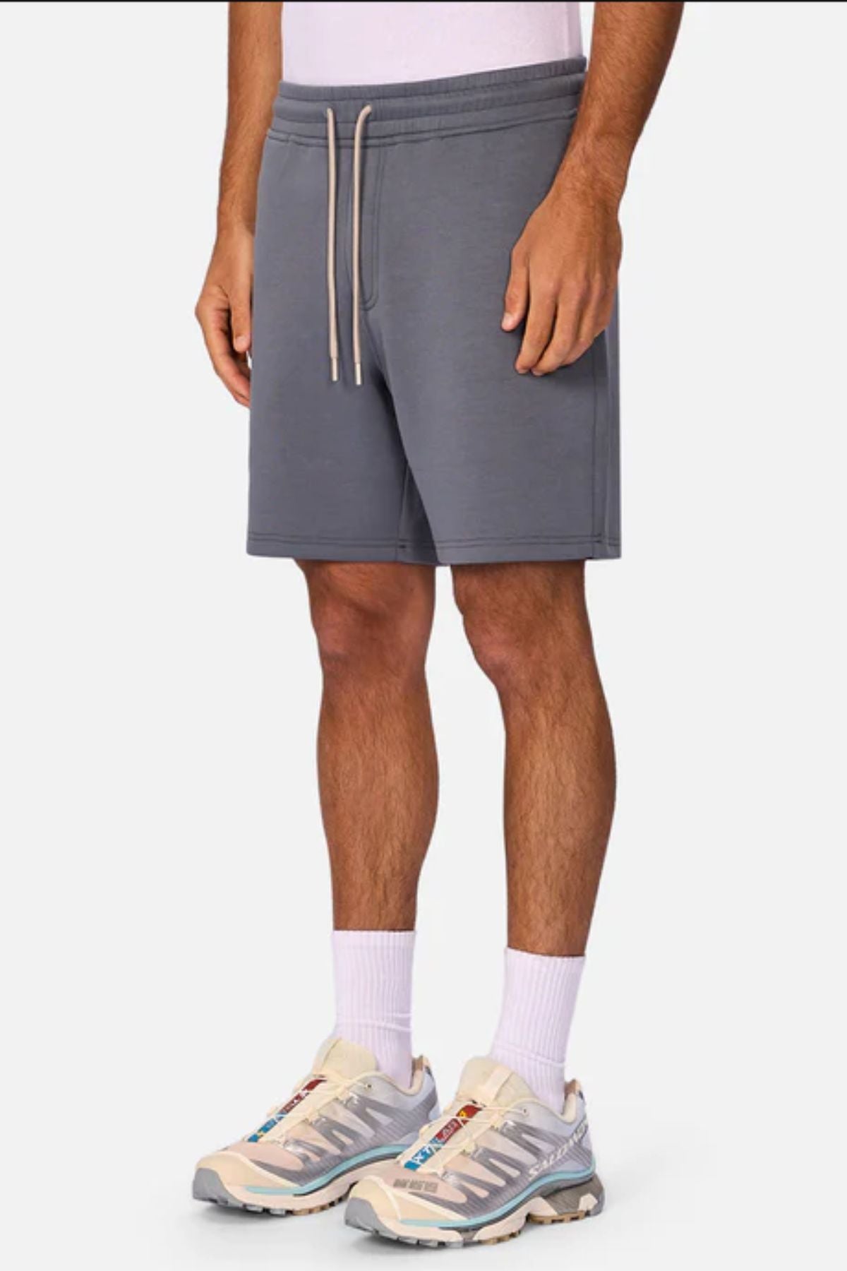 A person wears Industrie The Tech Track Short in gray, paired with a white shirt, white crew socks, and light sneakers, standing against a plain background. The image shows their look cropped from shoulders to feet.