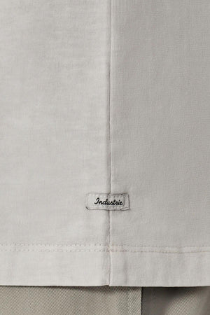 Close-up of the lower part of the Industrie The Del Sur Tee in light grey, featuring an oversized fit and a small stitched "Industrie" label near the hem. Grey pants are visible below this 100% cotton shirt.