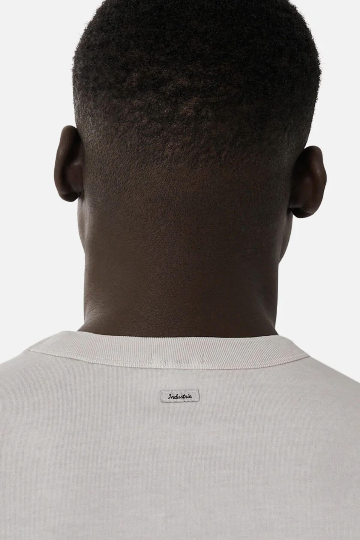 A person with short, closely-cropped hair is seen from behind wearing a light-colored Industrie The Del Sur Tee featuring a small rectangular tag at the back neckline, set against a plain, light background.