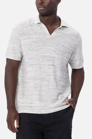 A modern man radiates confidence in a stylish polo, donning a light gray, short-sleeve Industrie The Lorenzo Polo with dark pants. He stands casually with one hand in his pocket against a white backdrop, gazing to the side.