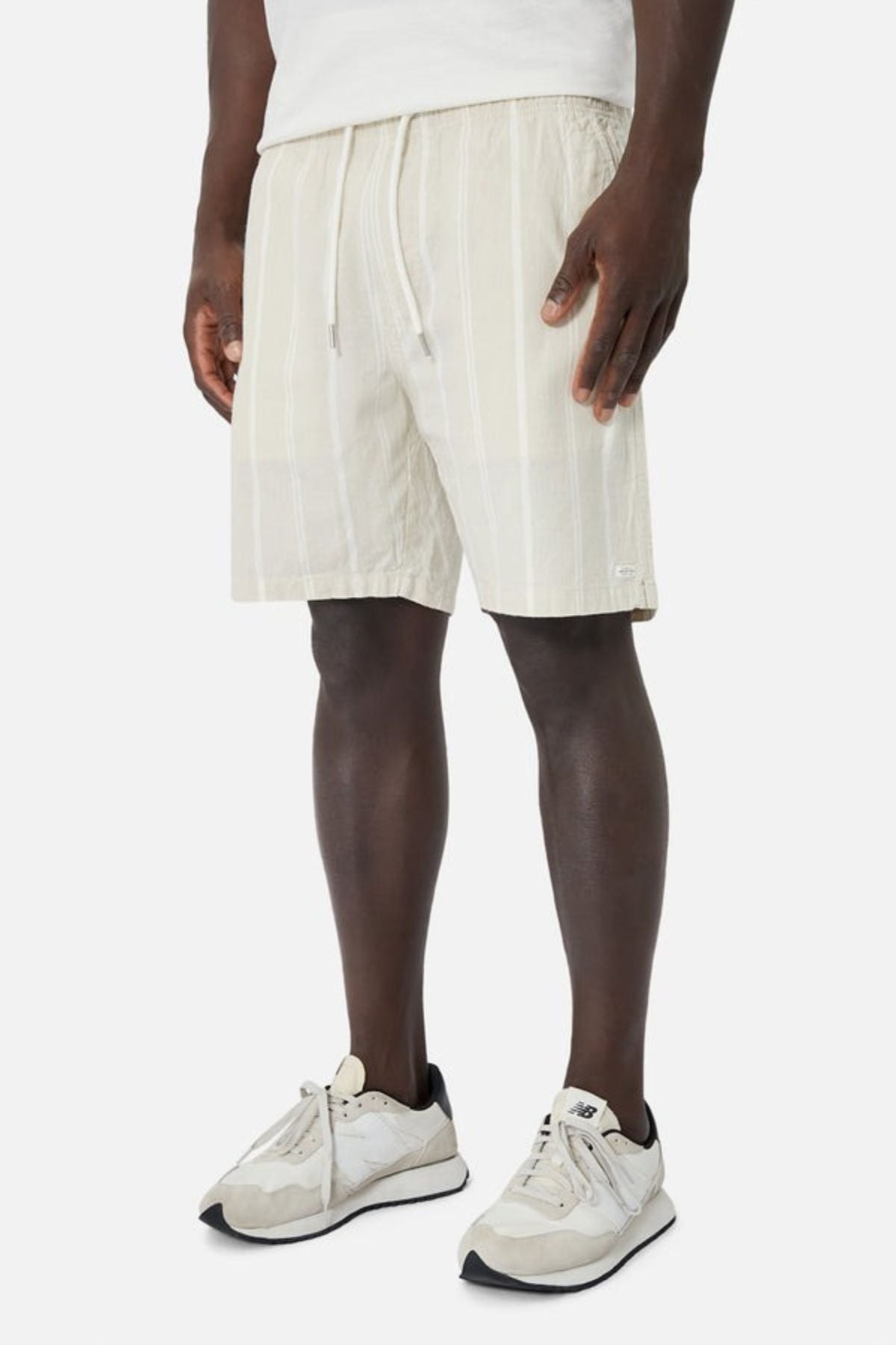 A person dressed in light-colored Industrie The Bel Air Shorts with an elastic waistband and a white top stands against a plain background. Complementing their casual wardrobe, they sport white sneakers with gray accents. The shorts feature subtle vertical stripes, adding a touch of flair.