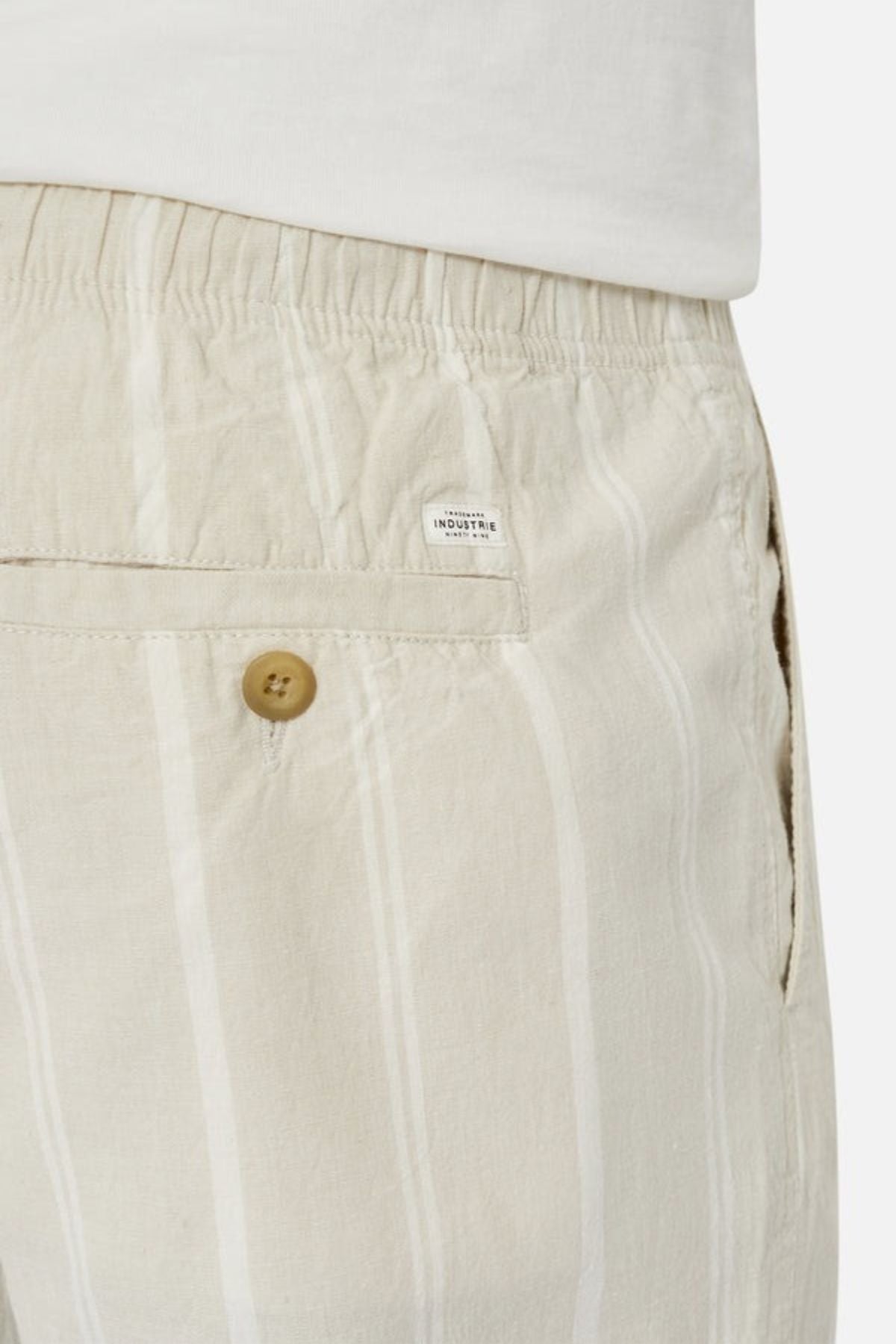 Close-up of a person wearing Industrie The Bel Air Short, showcasing beige and white stripes, an elastic waistband, and a buttoned back pocket. A small label sewn above the pocket adds a stylish touch to this casual wardrobe essential. The person completes the look with a crisp white shirt.