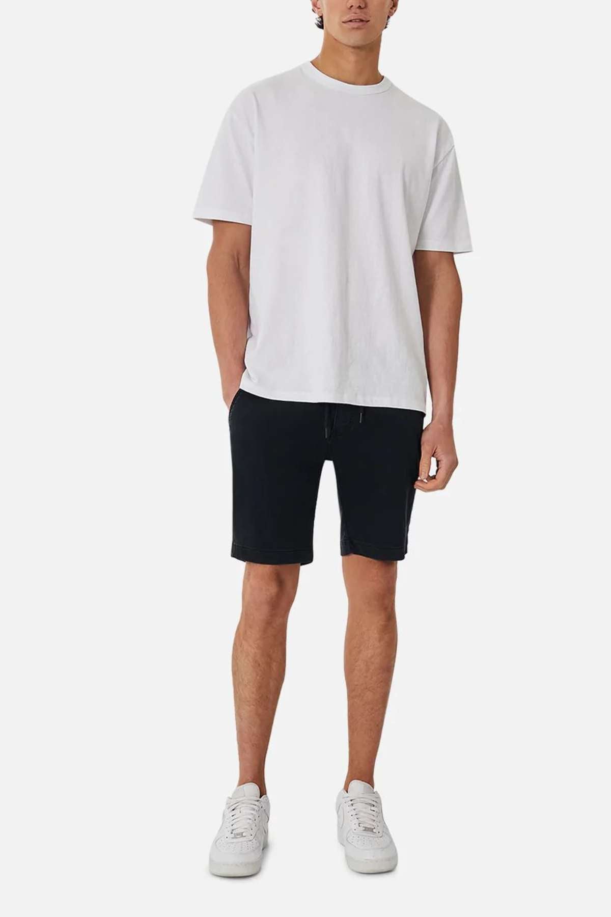 A person is standing against a plain white background, wearing a white t-shirt, Industrie The New Drifter Cuba Shorts with a comfortable belt loop waist, and white sneakers. Their face is not fully visible.