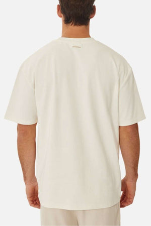 A person stands showcasing the oversized fit of Industrie's The Newport Tee in heavyweight fabric, paired with beige pants. The plain white background highlights the simplicity and style of their outfit.