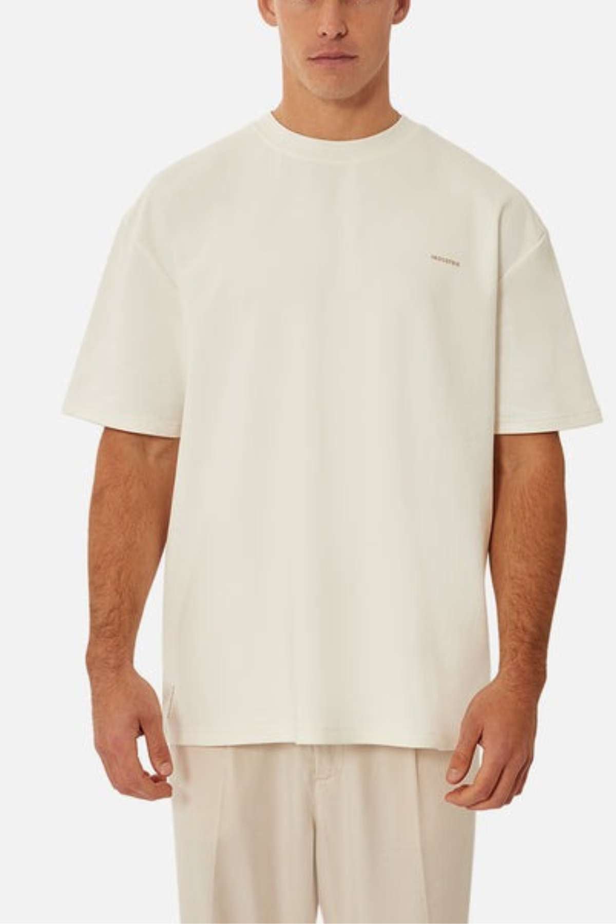 Against a plain background, a person wears Industrie’s The Newport Tee, made of heavyweight fabric. It has an oversized fit and short sleeves with minimal design. Their hands rest by their sides, capturing the relaxed vibe of this monochromatic outfit.