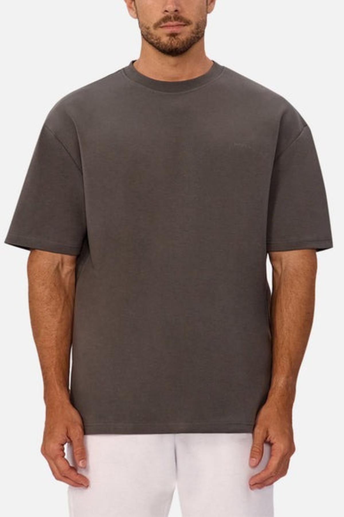 A man wearing a loose-fitting, short-sleeved Industrie The Newport Tee in heavyweight fabric and white pants stands facing forward against a plain light background.