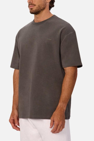 A man wears an oversized, dark gray Industrie The Newport Tee with subtle chest text and heavyweight fabric, paired with white pants, standing against a plain light background.