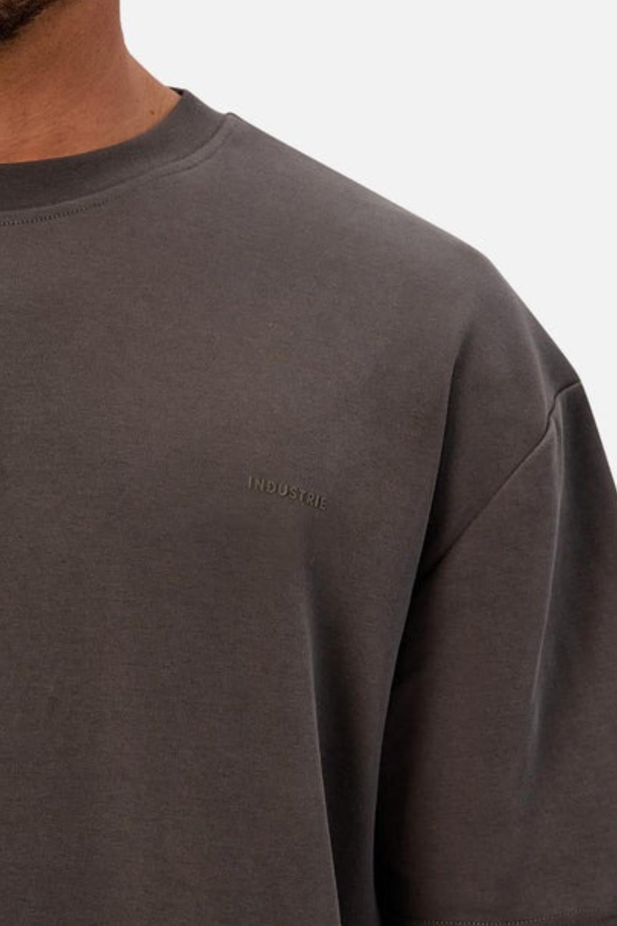 A close-up shows a person wearing the Industrie The Newport Tee, a plain, charcoal gray heavyweight crewneck with "INDUSTRIE" subtly printed on the chest. Only part of their face, shoulder, and upper torso are visible.