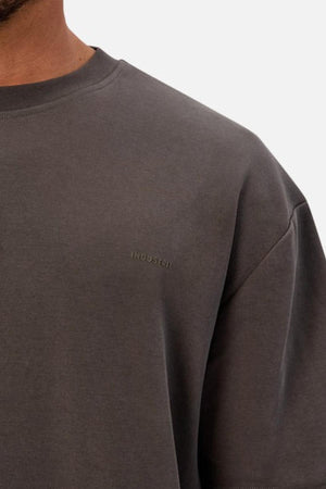 A close-up shows a person wearing the Industrie The Newport Tee, a plain, charcoal gray heavyweight crewneck with "INDUSTRIE" subtly printed on the chest. Only part of their face, shoulder, and upper torso are visible.
