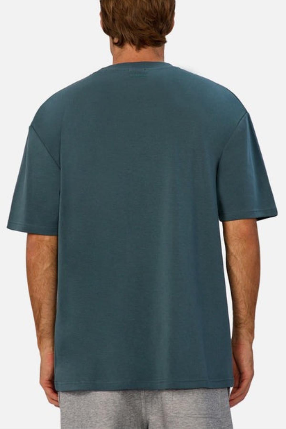 A person with light brown hair is shown from behind, wearing the Industrie The Newport Tee in teal and light gray shorts—ideal for a casual wardrobe—against a plain light background.