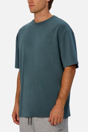 A man stands against a plain light background, wearing Industrie The Newport Tee in oversized teal and light gray sweatpants, with his arms relaxed by his sides.