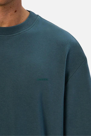 A close-up shows a person wearing the Industrie The Newport Tee, a teal crew neck made from heavyweight fabric with subtle "INDUSTRIE" embroidery on the chest, set against a plain light background.