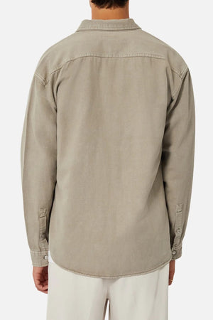 A person is seen from the back wearing Industrie The Portland Shirt, a relaxed-fit light gray denim jacket with buttoned cuffs, resembling an overshirt. It’s paired with cream-colored trousers for a casual look against a plain background.