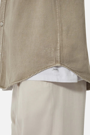 A close-up of a person wearing Industrie The Portland Shirt, a relaxed-fit beige overshirt made from a cotton linen blend, over a white T-shirt. Both items feature subtle branding near the hem for an understated look and are paired with light-colored pants.