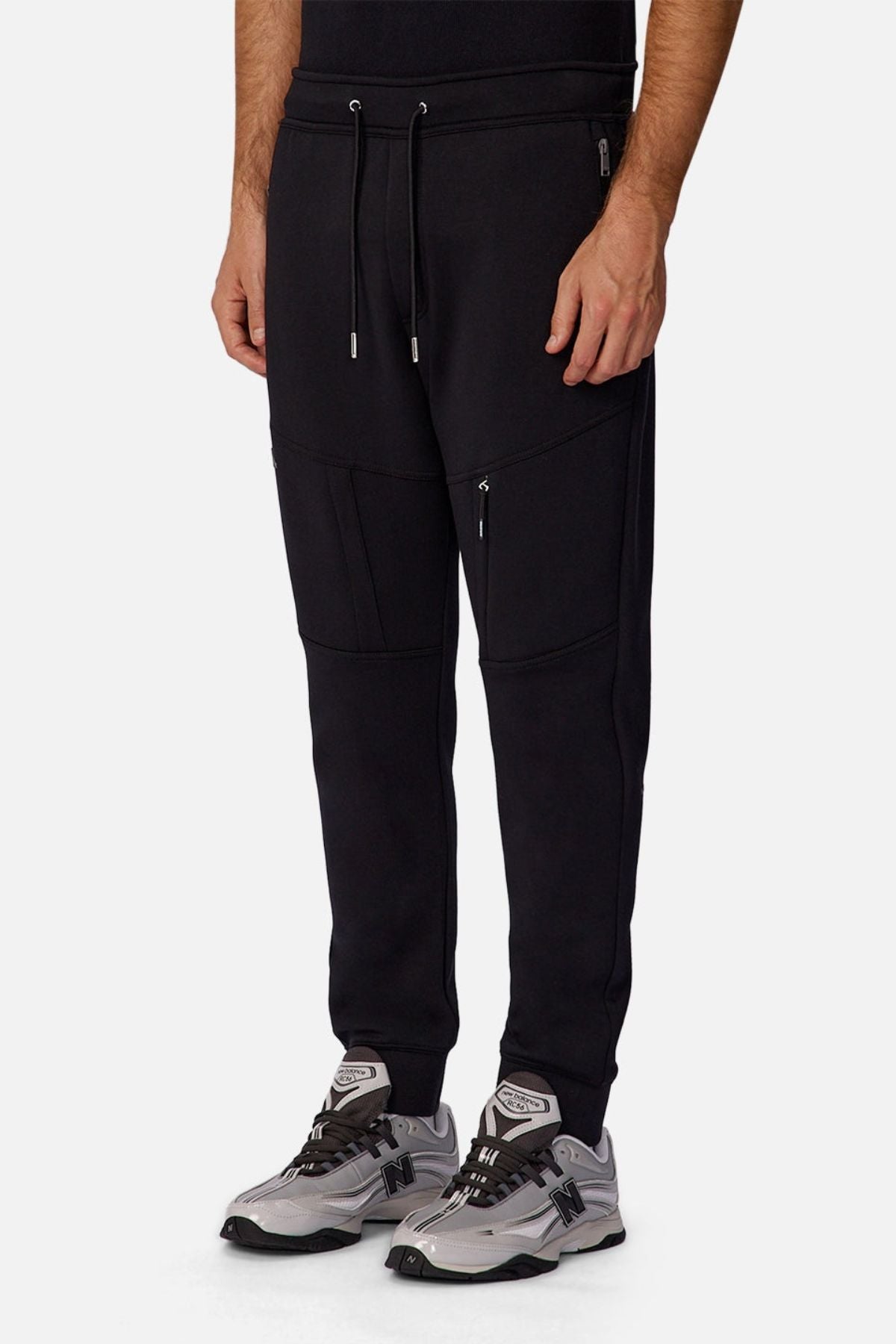 Wearing Industrie The Tech Armoured Track Pant joggers with combat pockets and gray athletic sneakers, a person stands against a white background. Only the lower half is shown, highlighting the pants’ tactical style.