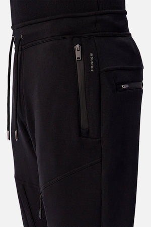 Close-up of a person wearing Industrie The Tech Armoured Track Pant in black, featuring zippered combat pockets, drawstrings, modern seams, and a small INDUSTRIE label by the pocket for a sleek, tactical style.