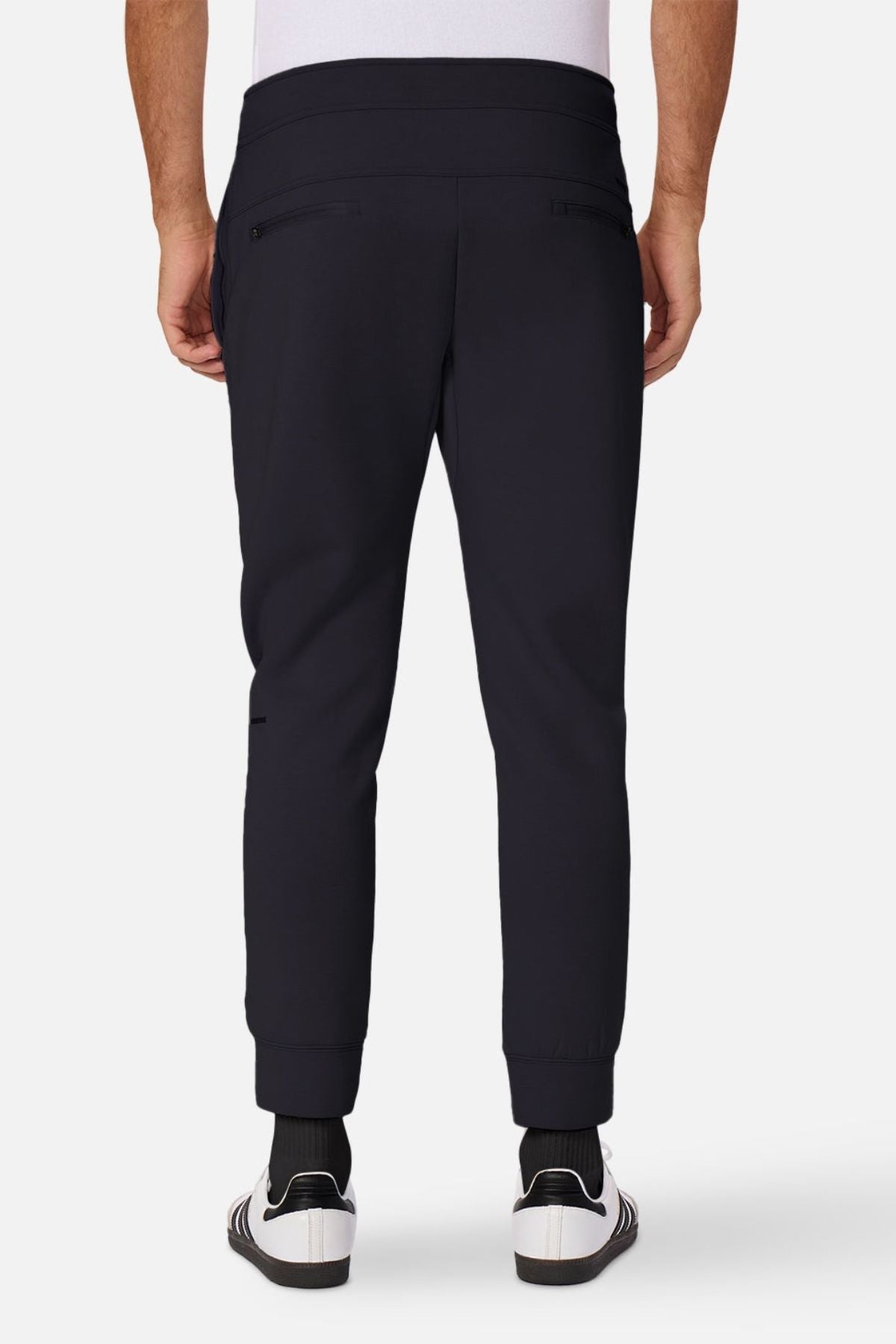 Someone is seen from behind wearing Industrie The Tech Armoured Track Pant in black with combat pockets, paired with white socks and white sneakers with black stripes, against a plain white background.