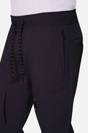 A close-up of a person wearing Industrie The Tech Armoured Track Pant in black, featuring zippered and combat pockets, a drawstring waistband, and a white shirt tucked in. The athletic fabric provides comfort and modern style.