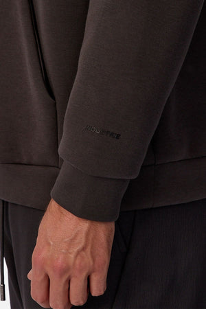 A close-up of a person's left arm and hand wearing the Industrie The Tech Del Sur Hoodie, featuring "INDUSTRIE" subtly printed near the wrist. The hand rests casually by their side, with matching dark pants partially visible.