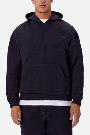A man stands against a white background wearing the Industrie The Tech Del Sur Hoodie, a plain black pullover with a front pocket and hood down, revealing a white T-shirt underneath, paired with matching black pants.