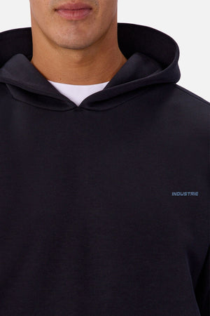 A close-up of a person in the Industrie The Tech Del Sur Hoodie, a black athletic pullover with a small "INDUSTRIE" logo on the left chest, layered over a white t-shirt. The person's face is partially visible.