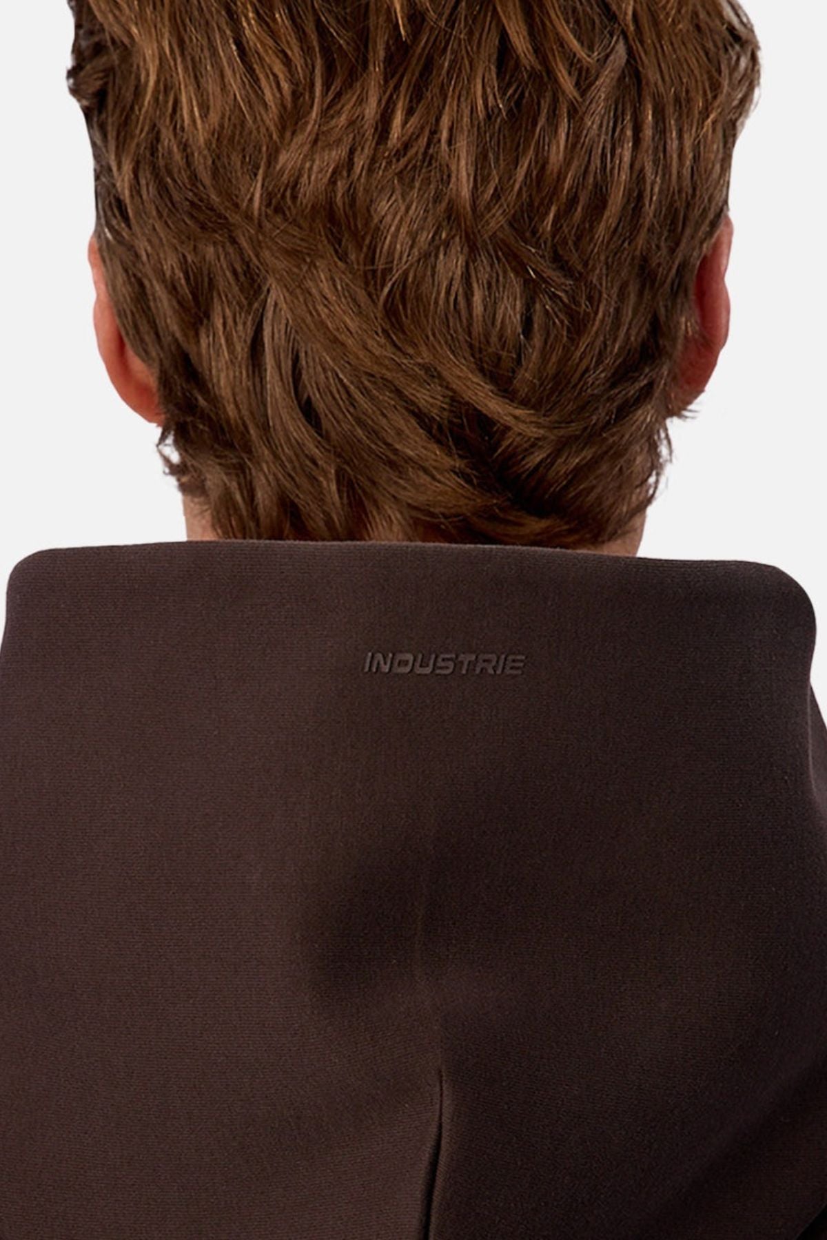 A close-up of the back of a person’s head and upper shoulders showcases the Industrie The Tech Del Sur Hoodie in dark brown, featuring "INDUSTRIE" subtly embroidered on the collar. Breathable fabric pairs with their light brown, wavy hair.