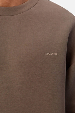 A close-up of a person wearing Industrie The Tech Del Sur Sweat in brown, featuring small white "INDUSTRIE" embroidery on the left chest. The crew-neck sweatshirt is designed for active lifestyles; the person's face is not shown.