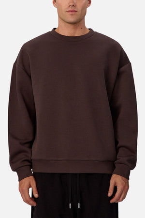 A man wears the Industrie The Tech Del Sur Sweat, a loose-fitting, dark brown crew neck with long sleeves, paired with black pants, standing against a plain light background.