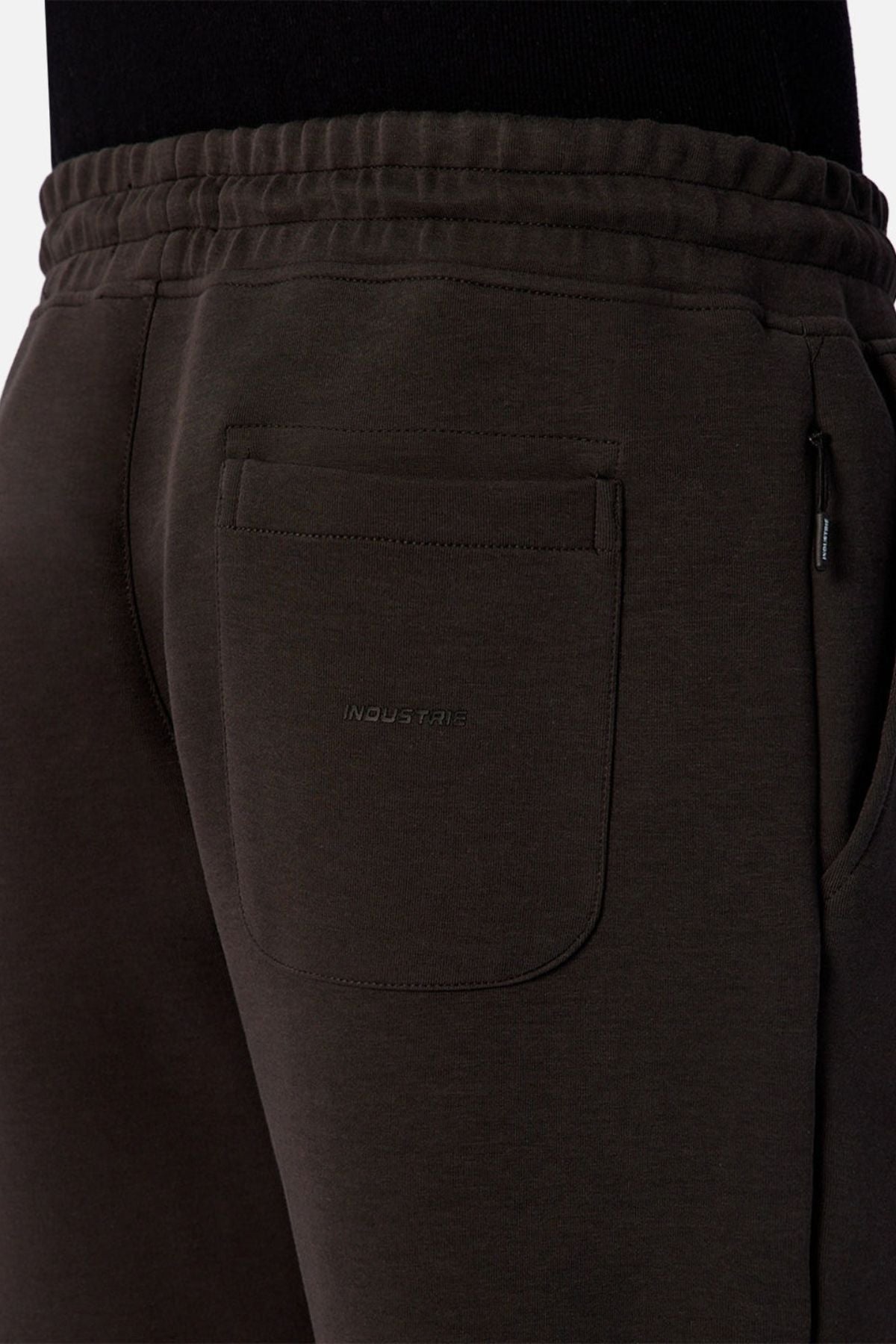 Close-up of the back pocket area of dark brown Industrie The Tech Del Sur Track Pant made from athletic material, featuring an elastic waistband and subtle "INDUSTRIE" embroidery on the pocket. The wearer pairs it with a black top.
