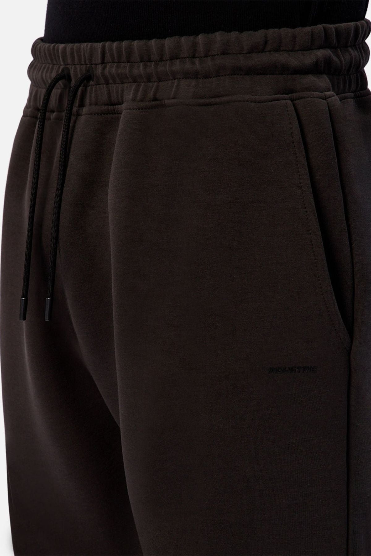 Close-up of a person wearing Industrie The Tech Del Sur Track Pant—dark brown joggers with an elastic waistband, drawstrings, and side pocket. Made from soft athletic material and styled with a tucked black top for a comfortable look.