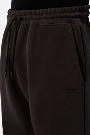 Close-up of a person wearing Industrie The Tech Del Sur Track Pant—dark brown joggers with an elastic waistband, drawstrings, and side pocket. Made from soft athletic material and styled with a tucked black top for a comfortable look.