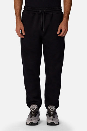 A person models the Industrie The Tech Del Sur Track Pant in black with a drawstring waist, paired with gray athletic shoes against a plain white background. The image is cropped from the chest down.