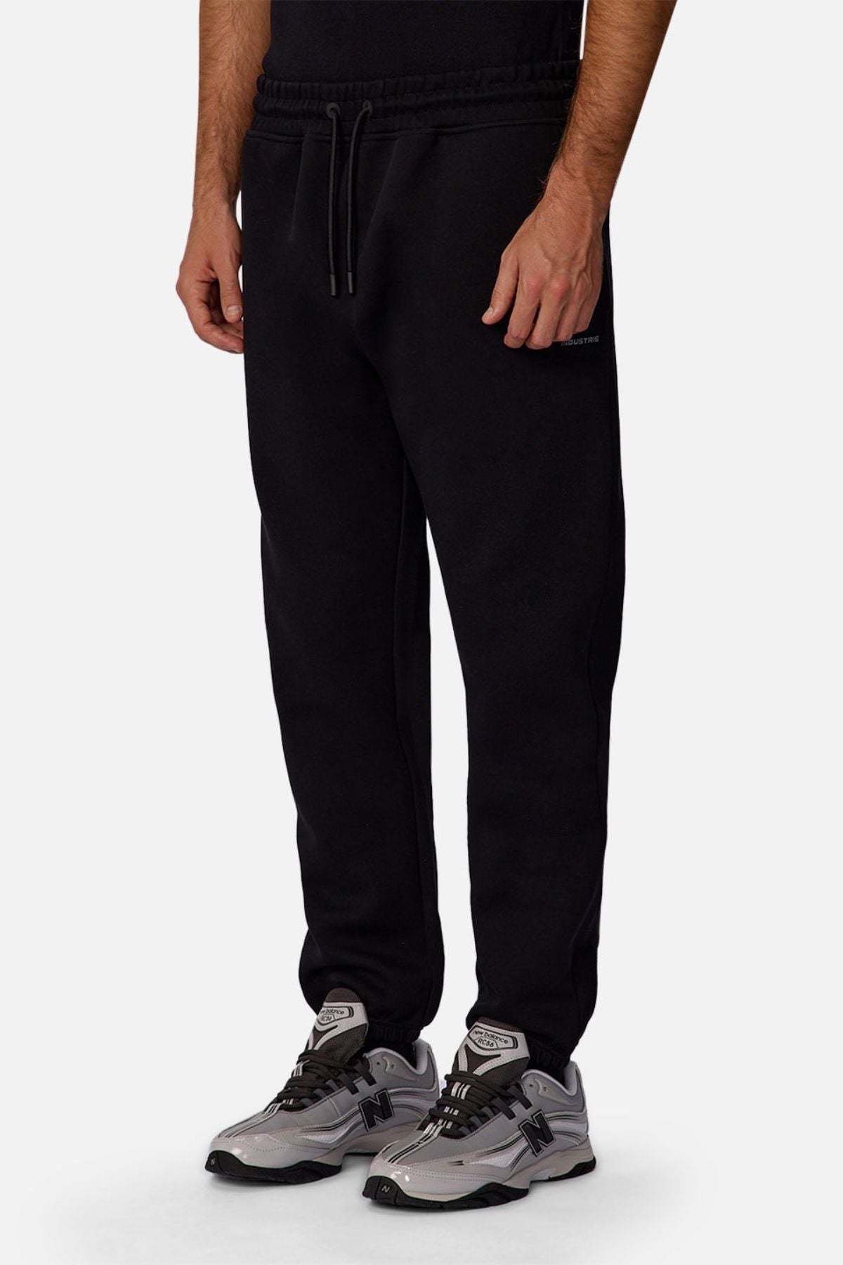 Someone wearing Industrie The Tech Del Sur Track Pant jogger sweatpants with a drawstring waist and gray New Balance sneakers stands against a plain white background, with only the lower body visible.