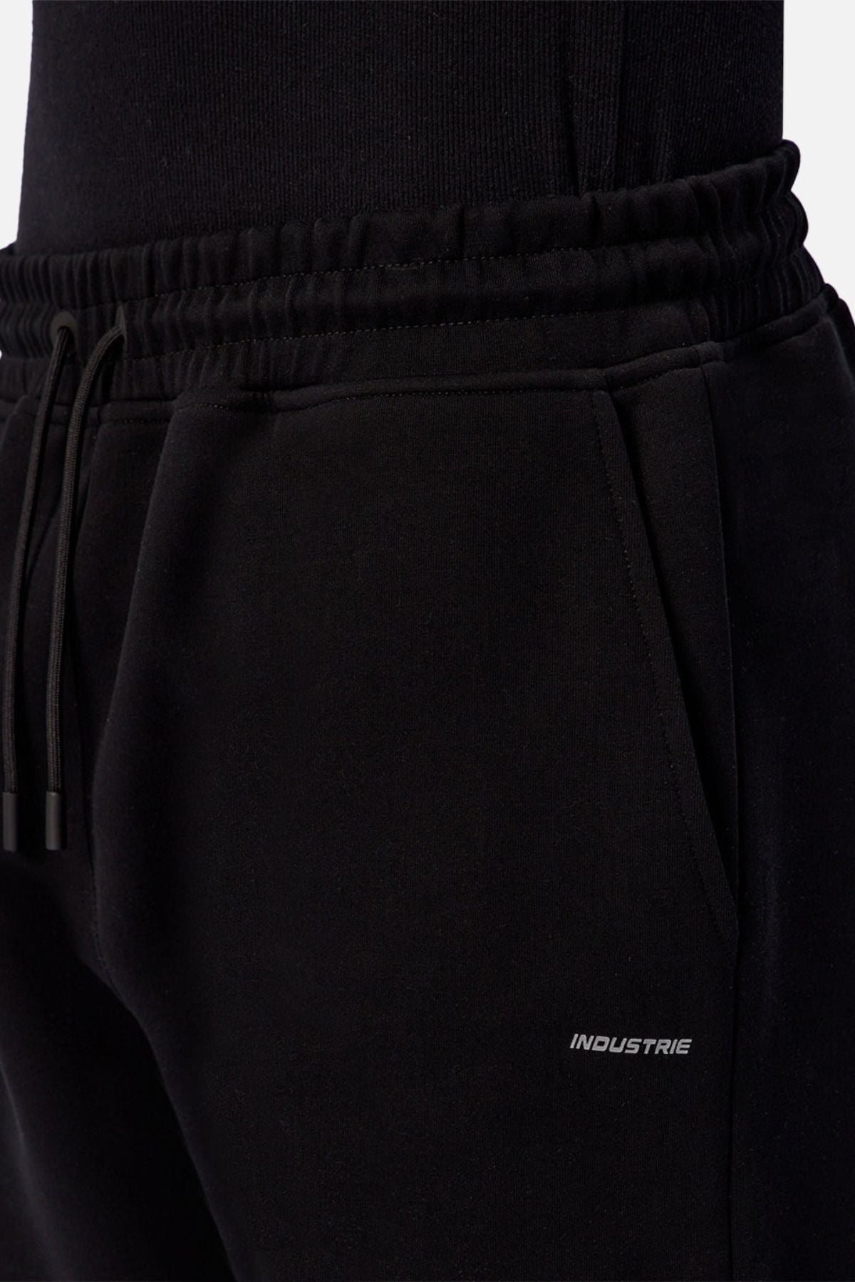 A close-up of a person wearing Industrie The Tech Del Sur Track Pant in black, featuring an elastic waistband with a drawstring and a small "INDUSTRIE" logo printed near the right pocket.