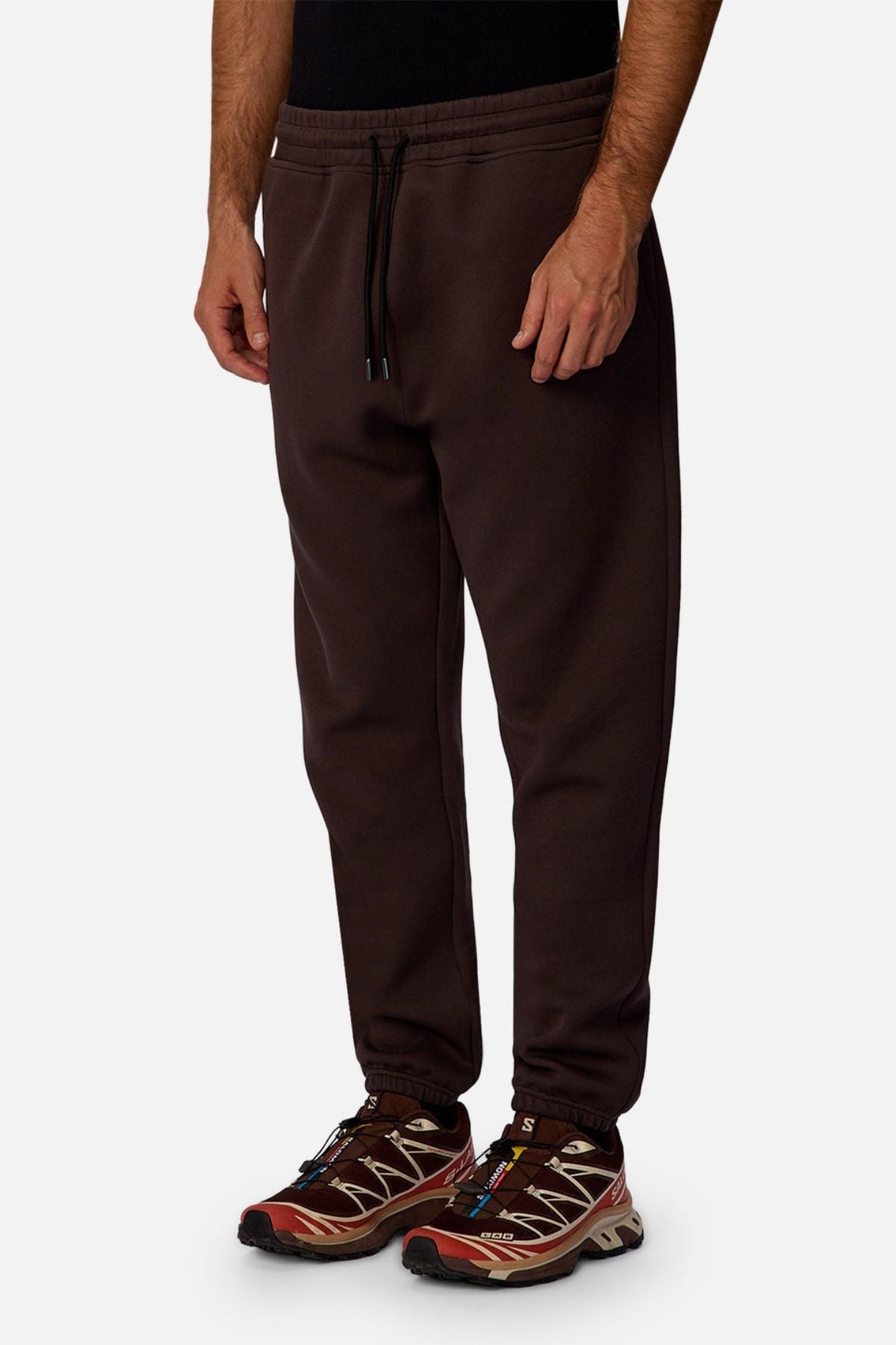 A person wears the Industrie The Tech Del Sur Track Pant in dark brown with a drawstring waist, paired with red and brown sneakers, standing against a plain white background; the upper body is partially out of frame.