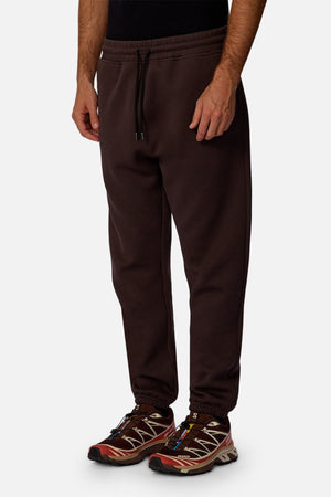 A person wears the Industrie The Tech Del Sur Track Pant in dark brown with a drawstring waist, paired with red and brown sneakers, standing against a plain white background; the upper body is partially out of frame.