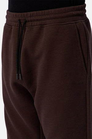 A close-up of a person wearing the Industrie The Tech Del Sur Track Pant in dark brown, featuring a black drawstring, black shirt tucked in, and subtle embossed Industrie branding near the pocket for a refined athletic look.