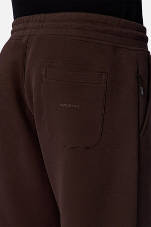 Close-up of a person wearing the Industrie The Tech Del Sur Track Pant in dark brown, featuring embroidered branding above the back pocket. The athletic material, waistband, and side pocket are partially visible.