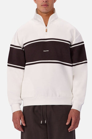A man wearing the Industrie The Tech Southend Track Top, a white quarter-zip sweatshirt with a black stripe and central white text, styled with dark pants for a casual, relaxed look.