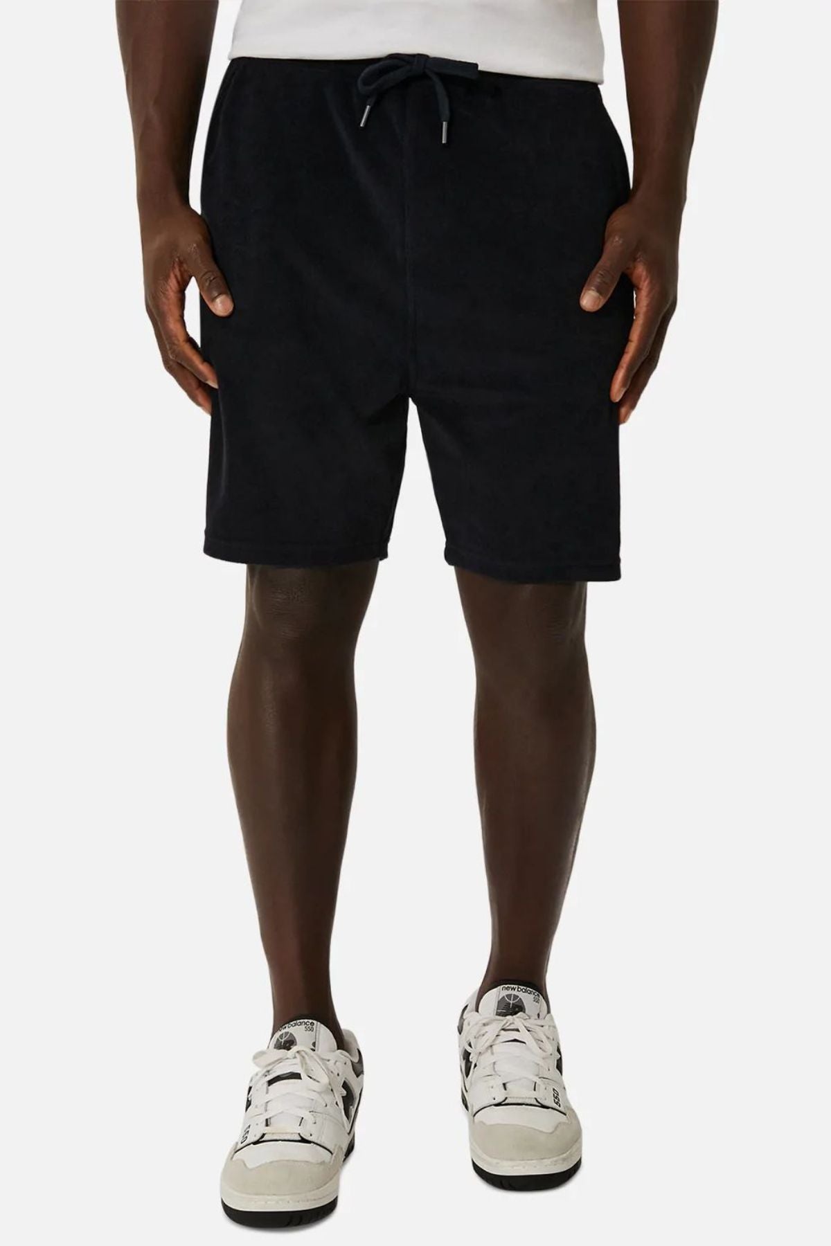 A person stands against a plain background wearing a white shirt and Industrie The Wynwood Short, perfect for a summer look. The black shorts feature a drawstring and are complemented by white sneakers with black accents. Their hands rest relaxed by their sides, embodying effortless style.