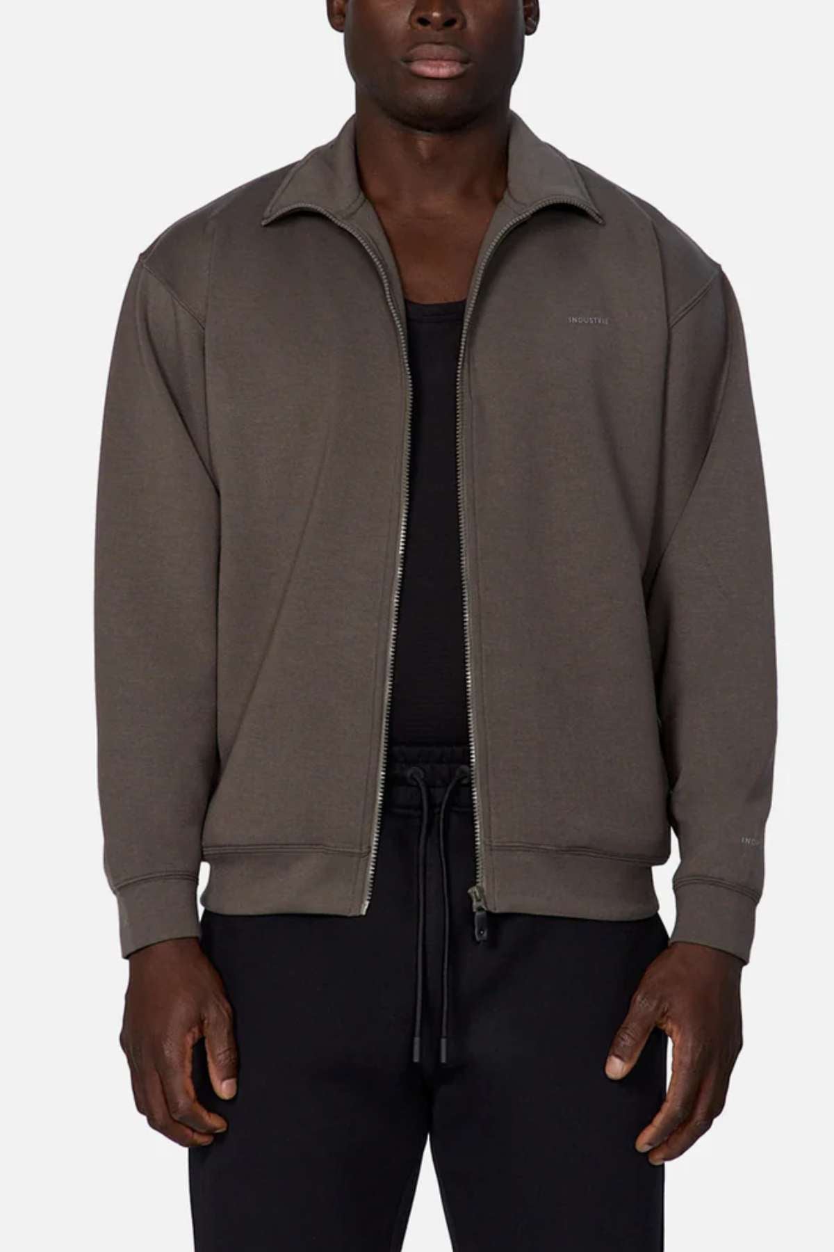 A man wears the Industrie The Zip Thru Track Top, made from soft dark gray fabric in an athletic style, layered over a black shirt and black pants, standing against a plain light background.