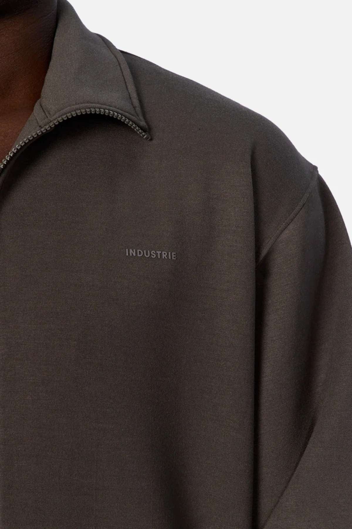 Close-up of someone wearing the Industrie The Zip Thru Track Top in dark grey, featuring soft fabric and "INDUSTRIE" embroidered on the chest. Only the shoulder and neck area show, emphasizing its athletic style.