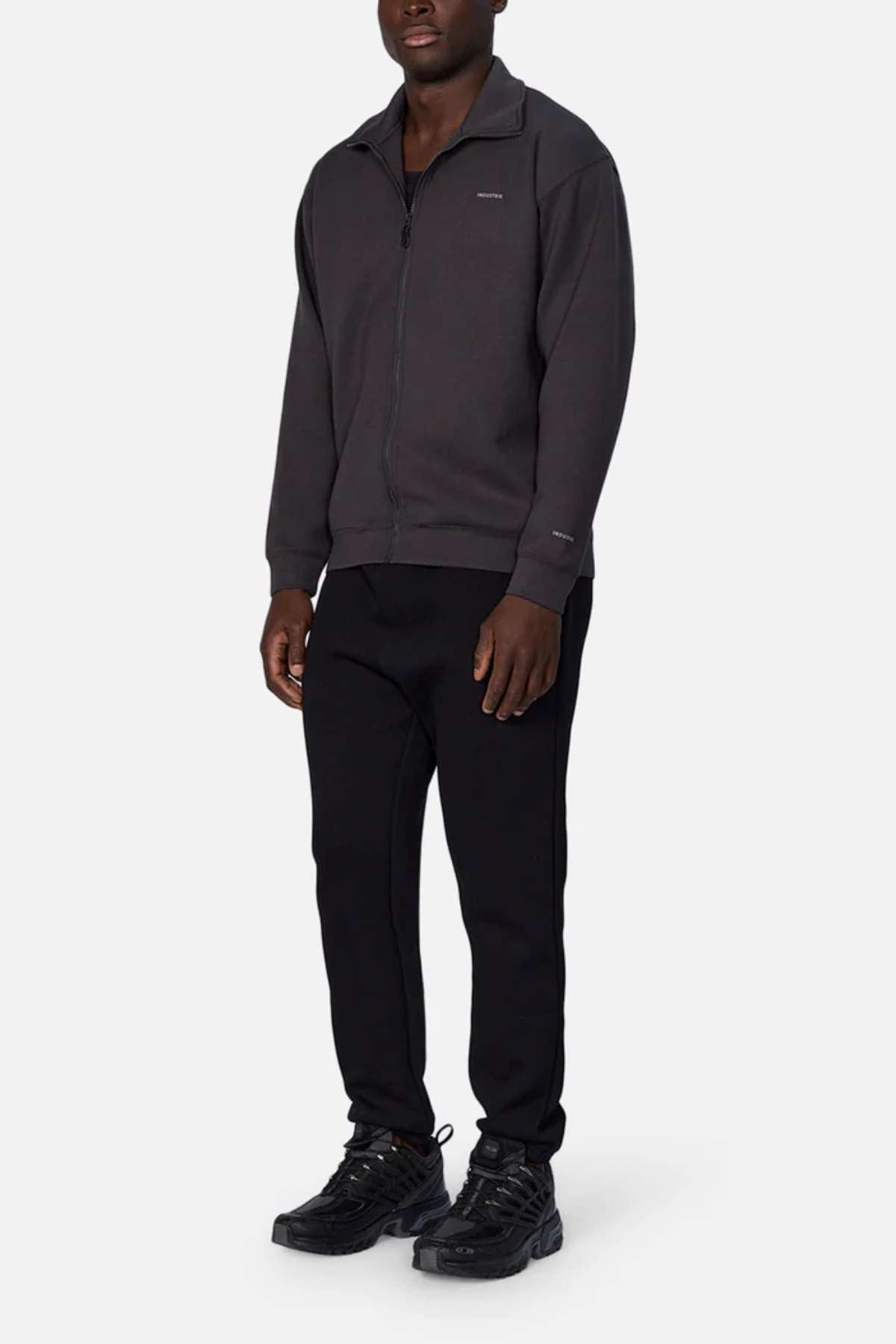 A person stands against a plain white background wearing the Industrie The Zip Thru Track Top, black pants, and black sneakers. The outfit is casual and monochromatic. The person has short hair and their hands rest by their sides, exuding an understated athletic style.