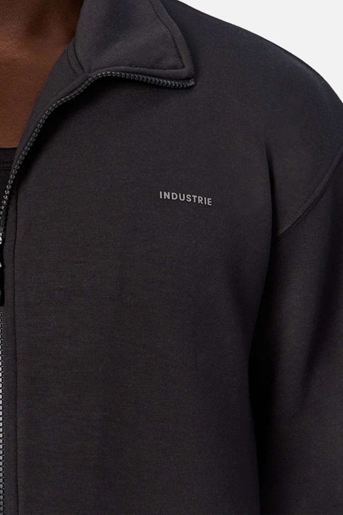 A close-up image of a person wearing the Industrie The Zip Thru Track Top jacket. The soft, trendy fabric enhances its athletic style, featuring the brand name "INDUSTRIE" written in small, white capital letters on the left side of the chest. The visible background is plain light gray.